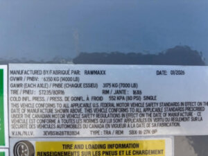 Close-up of a vehicle information label showing details such as manufacturer (RAWMAXX), date (01/2026), GVWR (6350 kg/14000 lb), GAWR (3175 kg/7000 lb), tire size, rim size, and safety compliance information. -Stillwater Trailer Sales