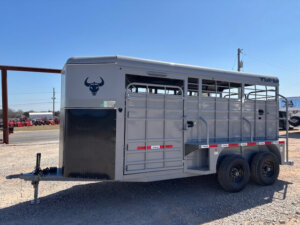 A gray livestock trailer with two axles and black wheels is parked on gravel. The trailer has a bull head logo, barred side panels, red and white reflective tape, and an open side door. Power lines and vehicles are in the background. -Stillwater Trailer Sales