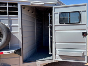 A gray Swift Built bumper pull stock trailer with its side door open reveals the empty interior and a spare tire mounted on the side. The 16' stock trailer features metal walls, a small window, and a textured floor. -Stillwater Trailer Sales