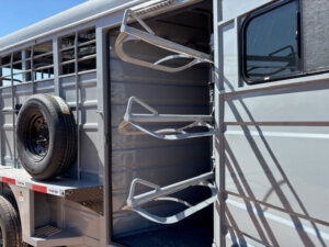 Open side door of a gray Swift Built bumper pull trailer reveals three empty saddle racks and a mounted spare tire on the exterior, with sunlight casting shadows on the stock trailer. -Stillwater Trailer Sales