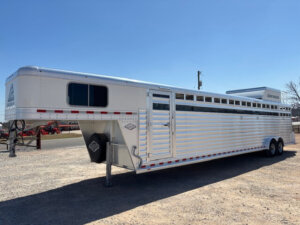 A large, silver Elite 10 Horse Trailer with multiple side vents is parked on gravel under a clear blue sky. The trailer has dual axles, a gooseneck hitch, and red and white reflective tape along the sides. -Stillwater Trailer Sales