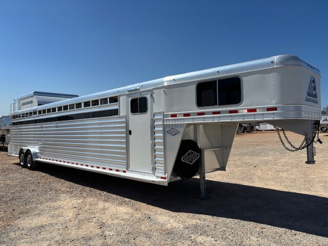 An Elite 10 Horse Trailer, large and silver with multiple axles and side vents, is parked on a gravel lot under a clear blue sky. This trailer features a gooseneck hitch and a small door on the side. -Stillwater Trailer Sales