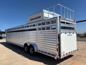 An Elite 10 Horse Trailer, large and silver with horizontal slats and a raised vented area on top, is parked on gravel under a clear blue sky. The trailer features dual wheels and a rear access door. -Stillwater Trailer Sales