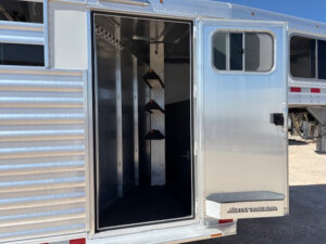 Open door of a shiny Elite 10 Horse Trailer reveals an interior tack room with three saddle racks mounted vertically on a metal post and carpeted floor. Sunlight shines on the exterior, and another trailer is visible nearby. -Stillwater Trailer Sales