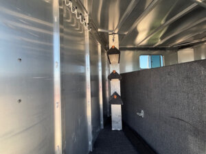 Interior of an Elite 10 Horse Trailer featuring three triangular, carpeted saddle racks mounted vertically on a support column; silver metal walls and ceiling with a dark carpeted floor offer durability and style. -Stillwater Trailer Sales