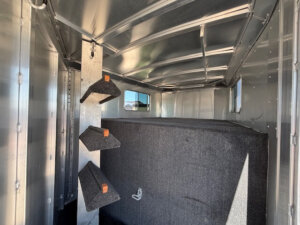 Interior of an Elite 10 Horse Trailer with three carpeted saddle racks mounted on the left wall and a large carpeted storage area on the right; sunlight shines through a window. -Stillwater Trailer Sales