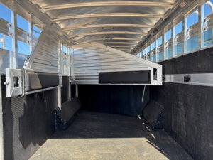 Interior view of an empty Elite 10 Horse Trailer with metal walls, a slatted roof, and a partially open metal gate dividing the space. Sunlight streams in through the roof slats, and the floor is covered with non-slip material. -Stillwater Trailer Sales
