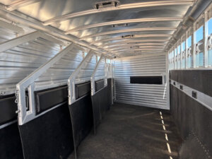 Interior of an empty Elite 10 Horse Trailer with metal walls, slatted windows along the right side, black partitions, and a non-slip floor. The trailer is well-lit, showing sturdy, clean construction. -Stillwater Trailer Sales