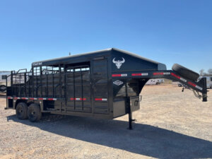 A black Swift Built livestock trailer with a gooseneck hitch is parked on a gravel lot under a clear blue sky. This 20' 3/4 Top model features tack boxes, a white bull head logo, and a mounted spare tire at the front. -Stillwater Trailer Sales