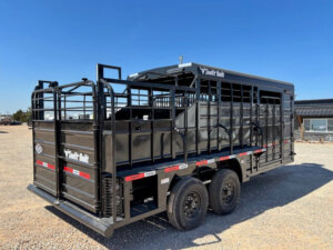 A black Swift Built livestock trailer with dual axles and tack boxes is parked on gravel under a clear blue sky. The 20' 3/4 top trailer features side rails, a rear gate, and branding. A building is visible in the background. -Stillwater Trailer Sales