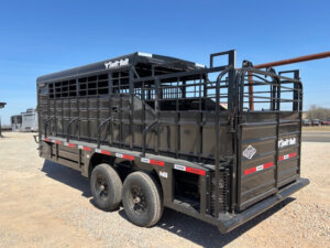 A black Swift Built livestock trailer with two axles, slatted sides for ventilation, tack boxes, and rear and side loading gates is parked on a gravel lot under a clear blue sky. This 20' 3/4 Top model offers both durability and convenience. -Stillwater Trailer Sales
