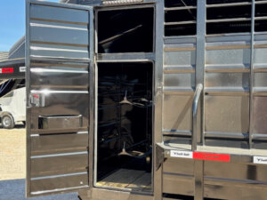 The image shows an open door on a shiny black Swift Built 20' livestock trailer, revealing an empty interior compartment with racks or shelves for Tack Boxes. Sunlight reflects off the 3/4 Top metal surface, with another vehicle visible in the background. -Stillwater Trailer Sales