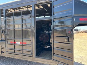 A black Swift Built livestock trailer with a 20' 3/4 Top and side door open reveals an interior section with tack boxes, a saddle rack, and wooden floor, parked on gravel under a clear sky. -Stillwater Trailer Sales