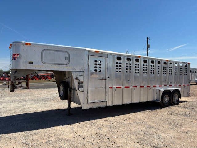 A large, silver Wilson livestock trailer with multiple vented windows is parked on a gravel lot under a clear, blue sky. The 24 foot stock trailer features two axles and a gooseneck hitch. -Stillwater Trailer Sales
