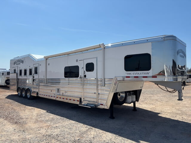 A large, shiny silver Bloomer gooseneck horse trailer with multiple windows, three axles, and spacious living quarters is parked on a gravel lot under a clear blue sky. "THE EVOLUTION" is written on the side. -Stillwater Trailer Sales