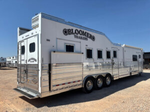 A large white Bloomer side load horse trailer with multiple windows and three axles is parked on a gravel lot under a clear blue sky. -Stillwater Trailer Sales