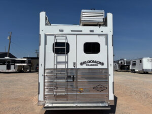 Rear view of a white Bloomer 5 Horse trailer with two windows, ladder on the left, air conditioning unit on the roof, and spacious living quarters, parked on a gravel lot with other trailers visible in the background. -Stillwater Trailer Sales