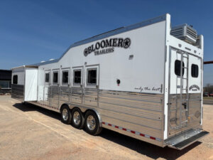 A large, silver and white Bloomer living quarters horse trailer with multiple windows and three axles is parked on a gravel lot under a clear blue sky. The trailer features the Bloomer logo and "only the best" slogan. -Stillwater Trailer Sales