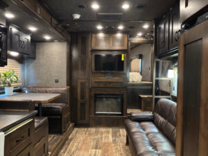 Interior of a modern RV living quarters with dark wood cabinetry, leather sofa, dining booth, flat-screen TV above an electric fireplace, potted plant on the table, and recessed ceiling lights&mdash;similar to a luxury Bloomer horse trailer setup. -Stillwater Trailer Sales