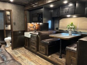 A cozy Bloomer RV interior featuring a kitchen with dark cabinets, farmhouse sink, microwave, dining area with a plant on the table, brown leather seating, wood-look flooring, and a view of the bathroom in the living quarters. -Stillwater Trailer Sales