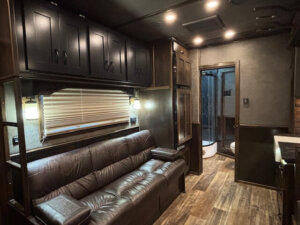 Interior of a modern horse trailer with stylish living quarters, featuring dark wood cabinetry, a brown leather sofa, wood-look flooring, and recessed lighting. A window with blinds sits above the sofa, and a bathroom with shower appears in the background. -Stillwater Trailer Sales