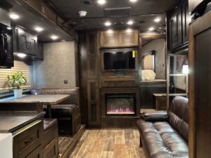 A cozy living quarters horse trailer interior featuring dark wood cabinets, a brown leather sofa, a dining nook with a potted plant, wall-mounted TV, and electric fireplace, with warm lighting and wood-style flooring. -Stillwater Trailer Sales