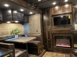 Interior of a 5 Horse Trailer with living quarters: dark wood cabinets, a built-in TV above an electric fireplace, leather dining booth, potted plant on the table, and wood floors. Warm, cozy lighting throughout this side load space. -Stillwater Trailer Sales