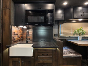 A modern RV kitchen in this Living Quarters Trailer features dark cabinets, a built-in microwave, farmhouse sink, brick backsplash, and a cozy dining area with a potted plant by the window with closed blinds. -Stillwater Trailer Sales