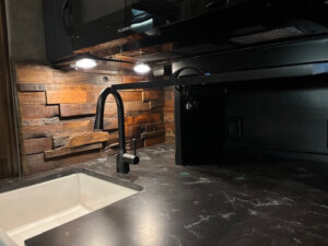 A modern kitchen countertop with a black marble surface, a white sink, matte black faucet, under-cabinet lighting, dark wooden backsplash, and a black microwave above&mdash;perfect for horse trailer living quarters with style and functionality. -Stillwater Trailer Sales