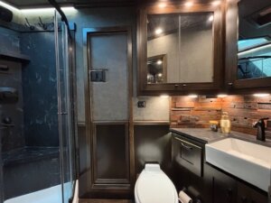 Modern bathroom in this Bloomer 5 Horse Trailer features dark wood cabinetry, a white toilet, rectangular sink, and shower with glass door. Soap and lotion dispensers sit on the counter. Warm lighting and textured walls create cozy living quarters. -Stillwater Trailer Sales