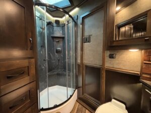 A compact bathroom in the 5 Horse Trailer Living Quarters features a glass-enclosed corner shower with dark walls, wooden cabinets, a toilet, and warm lighting. Sleek fixtures and brown and beige tones create a stylish, inviting space. -Stillwater Trailer Sales