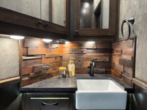 A modern bathroom vanity with a white rectangular sink, black faucet, and soap dispenser&mdash;perfect for a Living Quarters Horse Trailer. Rustic wooden planks form the backsplash, paired with dark wood cabinets and under-cabinet lighting. -Stillwater Trailer Sales