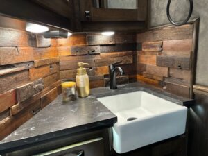 A rustic bathroom corner inside the living quarters of a 5 horse trailer features a white farmhouse sink, black faucet, marble countertop, wood plank backsplash, a gold soap dispenser, and a small candle&mdash; all illuminated by under-cabinet lighting. -Stillwater Trailer Sales