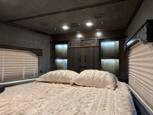 A cozy bedroom in the Bloomer 5 Horse Living Quarters features a neatly made bed with textured pillows and a cream coverlet, wood-paneled walls, sliding barn-style cabinet doors, and soft lighting from built-in shelves. Two windows have closed blinds. -Stillwater Trailer Sales