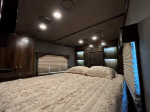 A cozy bedroom in a living quarters horse trailer features a large bed with a textured cream bedspread and two pillows, built-in cabinets with accent lighting above the bed, and a window with closed blinds on the left side. -Stillwater Trailer Sales
