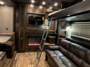 Interior of a 5 Horse Trailer featuring living quarters with a leather sofa, a fireplace with TV above, loft bed accessed by ladder, booth dining area, and wood cabinetry with a window covered by blinds. -Stillwater Trailer Sales