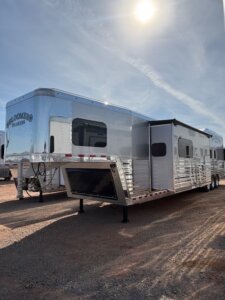 A shiny silver Bloomer 5 Horse Trailer with multiple windows is parked on a gravel lot under a bright sky, with other trailers visible in the background. -Stillwater Trailer Sales