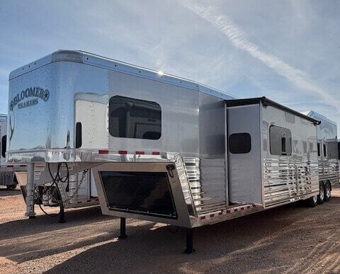 A shiny silver Bloomer horse trailer with slide-out living quarters is parked on gravel under a bright sun and blue sky. The trailer features multiple windows and reflective surfaces. -Stillwater Trailer Sales