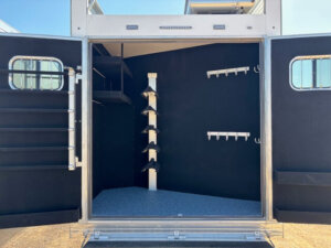 The image shows the open rear compartment of a 5 Horse Trailer with black interior walls, multiple hooks on the right, vertical saddle racks in the center, and shelving on the left. -Stillwater Trailer Sales