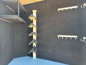 A corner of a horse trailer living quarters with black walls and blue speckled flooring features angled shelves on the left, a vertical metal rack with angled platforms in the center, and two rows of metal hooks and shelves on the right wall. -Stillwater Trailer Sales