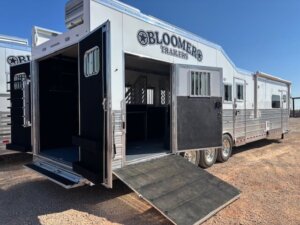 A large silver Bloomer 5 horse trailer with living quarters, a ramp, and open doors reveals spacious interior stalls. It&rsquo;s parked on gravel under a clear blue sky, showcasing its side load design for easy access. -Stillwater Trailer Sales