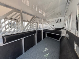 Interior of a clean, well-lit Bloomer horse trailer with padded dividers and metal bars creating separate stalls, featuring a textured floor and white walls. -Stillwater Trailer Sales