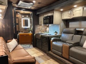 Modern RV interior in a Bloomer horse trailer featuring a brown leather sofa, two reclining chairs, a compact kitchen with sink, microwave, cabinets, and a vanity with mirror in the background&mdash;all warmly lit for luxurious living quarters. -Stillwater Trailer Sales