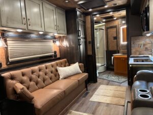 Interior of a luxury Bloomer RV 5 Horse Living Quarters with a tan tufted leather sofa, light-colored cabinets, a kitchenette, and a bathroom with shower. Warmly lit with elegant wood and leather accents throughout. -Stillwater Trailer Sales