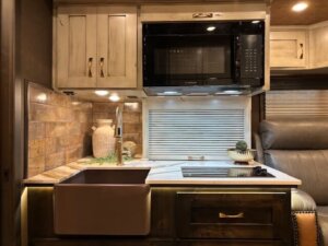 A modern RV kitchen in a Bloomer horse trailer features a farmhouse sink, white countertop, built-in stovetop, microwave, and decor like plants and vases&mdash;all in a warm, well-lit space with wood and tile accents. -Stillwater Trailer Sales