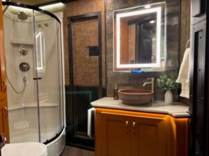 Modern bathroom inside a Bloomer horse trailer features a glass shower, illuminated mirror, brown vessel sink on a countertop, orange vanity cabinets, and stylish wall tiles&mdash;offering luxurious living quarters for your travels. -Stillwater Trailer Sales