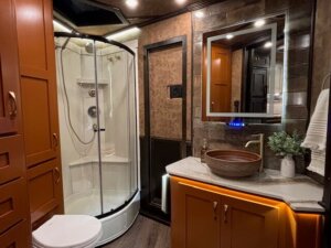 Modern bathroom with a curved glass shower, wooden cabinets, vessel sink on marble countertop, illuminated mirror, towel rack, and potted plant&mdash;like the comfort of living quarters in a Bloomer horse trailer&mdash;features warm lighting and brown-toned decor. -Stillwater Trailer Sales