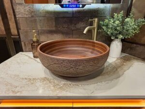 A modern bathroom sink in the living quarters of a 5 horse trailer features a round, textured wooden basin on a marble countertop. A gold faucet, soap dispenser, and a green leafy plant in a white vase complete the stylish look. -Stillwater Trailer Sales