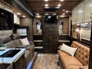 The interior of this Bloomer horse trailer features a leather sofa, reclining chairs, a small kitchen area, mounted TV, overhead cabinets, and windows with blinds&mdash;all with warm lighting and wooden floors for a cozy living quarters horse trailer atmosphere. -Stillwater Trailer Sales