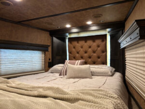 A cozy bedroom with a large, tufted headboard, neutral bedding, and decorative pillows serves as inviting living quarters inside this 5 horse trailer. Wood-paneled walls, recessed lighting, and side load windows create a warm retreat. -Stillwater Trailer Sales